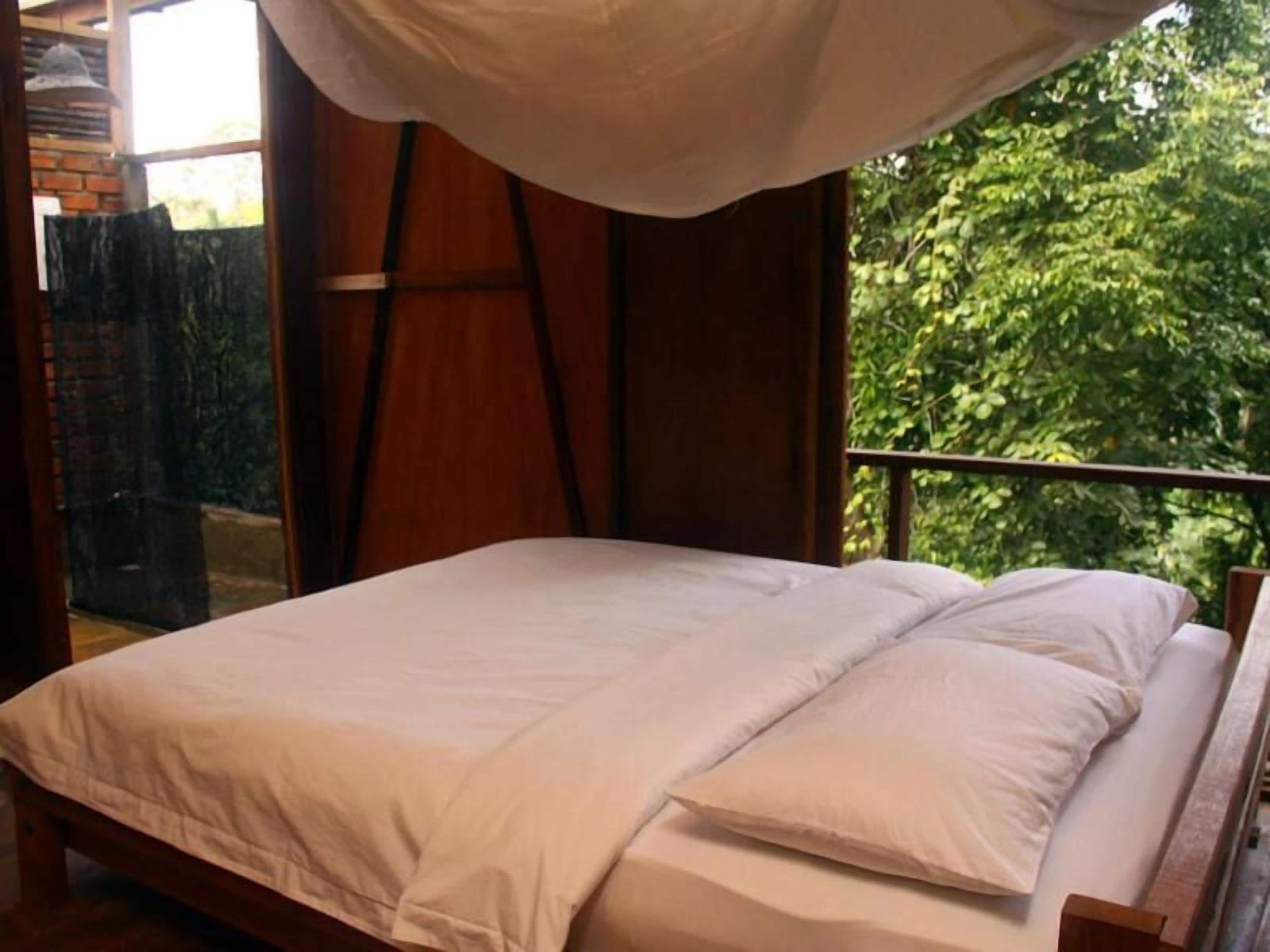 Een bed met witte lakens in een kamer met houten vloer en uitzicht op groen.