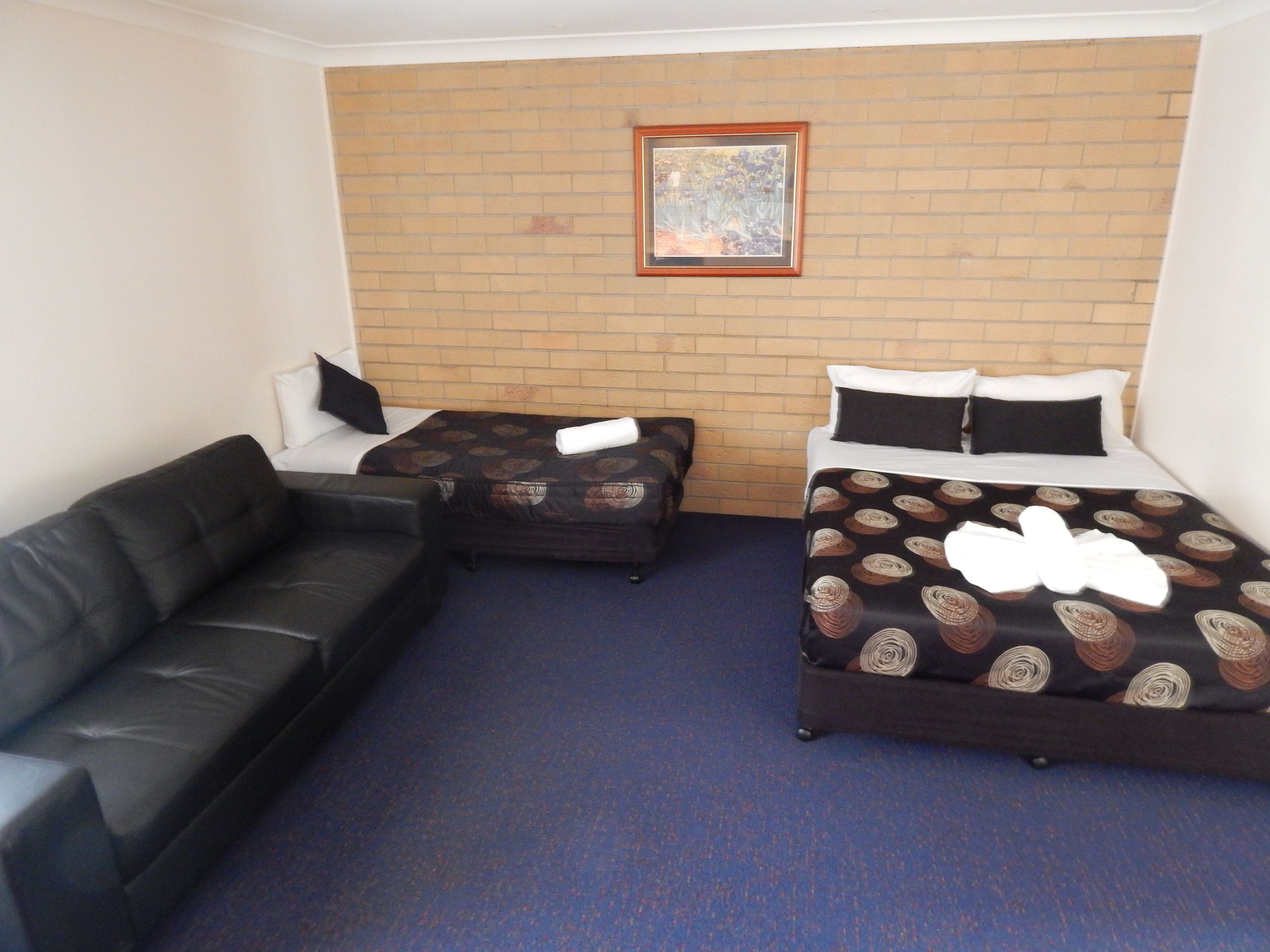 A hotel room with a brick wall, two single beds with patterned bedding, a black leather sofa, and a framed picture above the beds.
