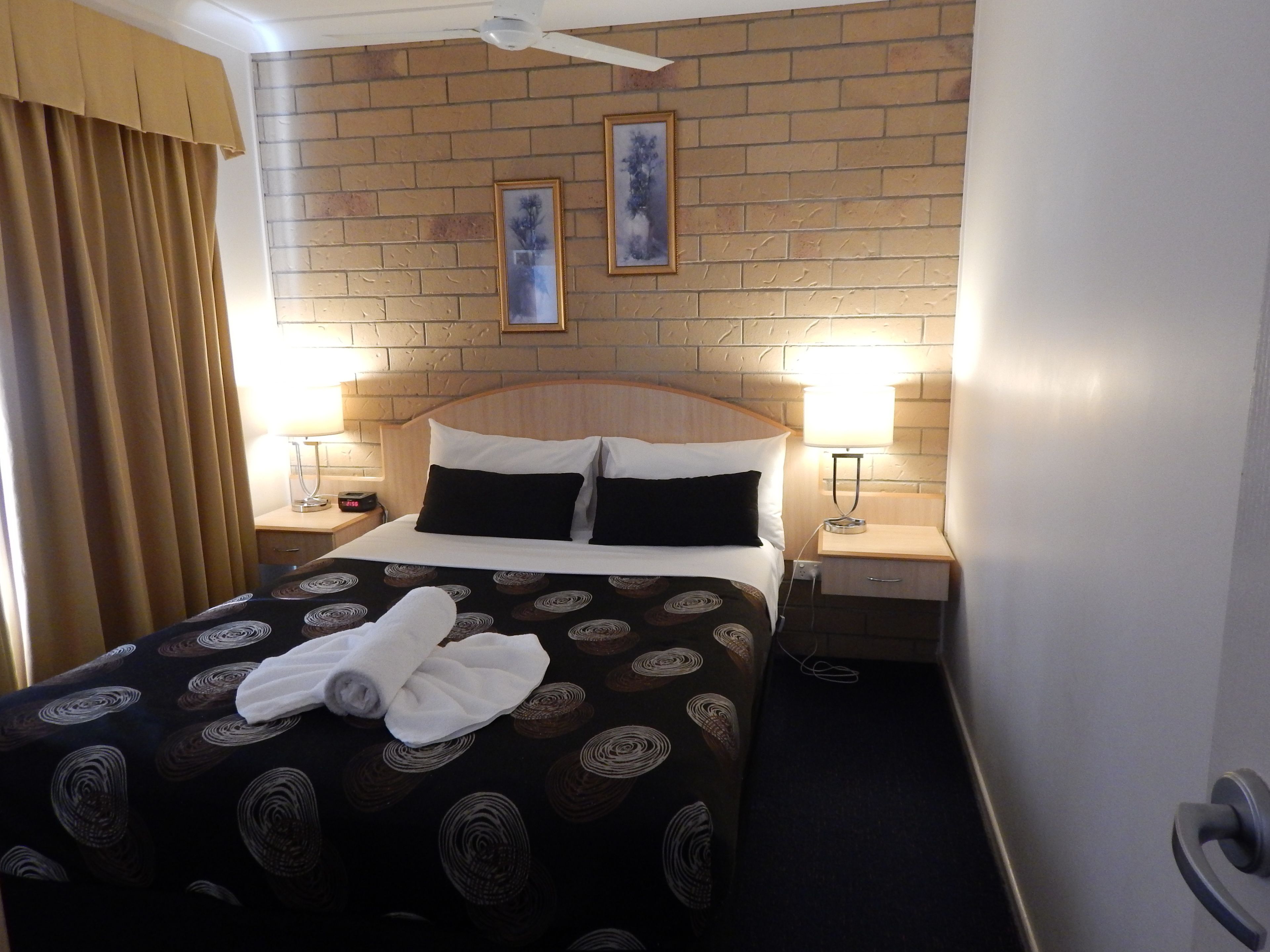 Une chambre d’hôtel avec une tête de lit décorée d’un motif en briques, deux lampes de chevet, un lit recouvert d’une couvre-lit à motifs et deux tableaux encadrés accrochés au mur.