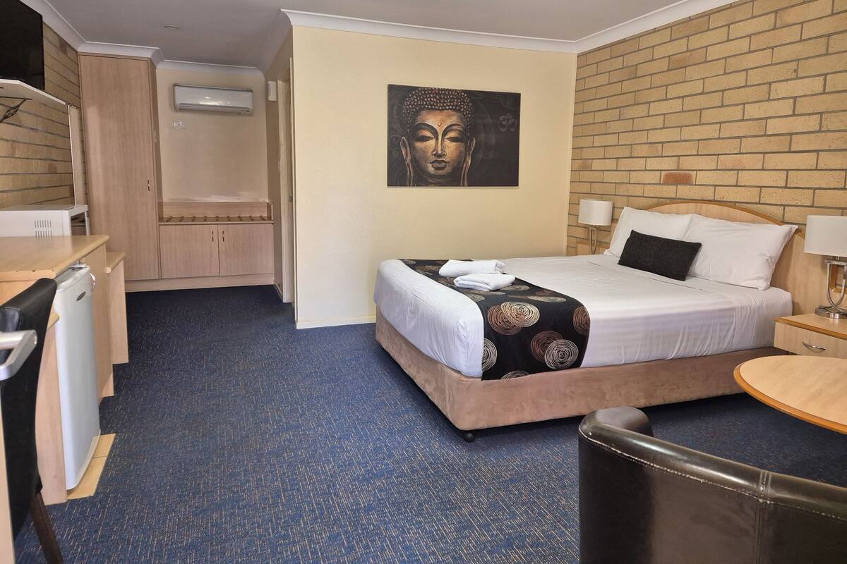 A hotel room with a bed, a desk, a chair, a small table, a wall-mounted air conditioner, and a painting on the wall.