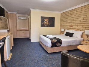 A hotel room with a bed, a desk, a chair, a small table, a wall-mounted air conditioner, and a painting on the wall.