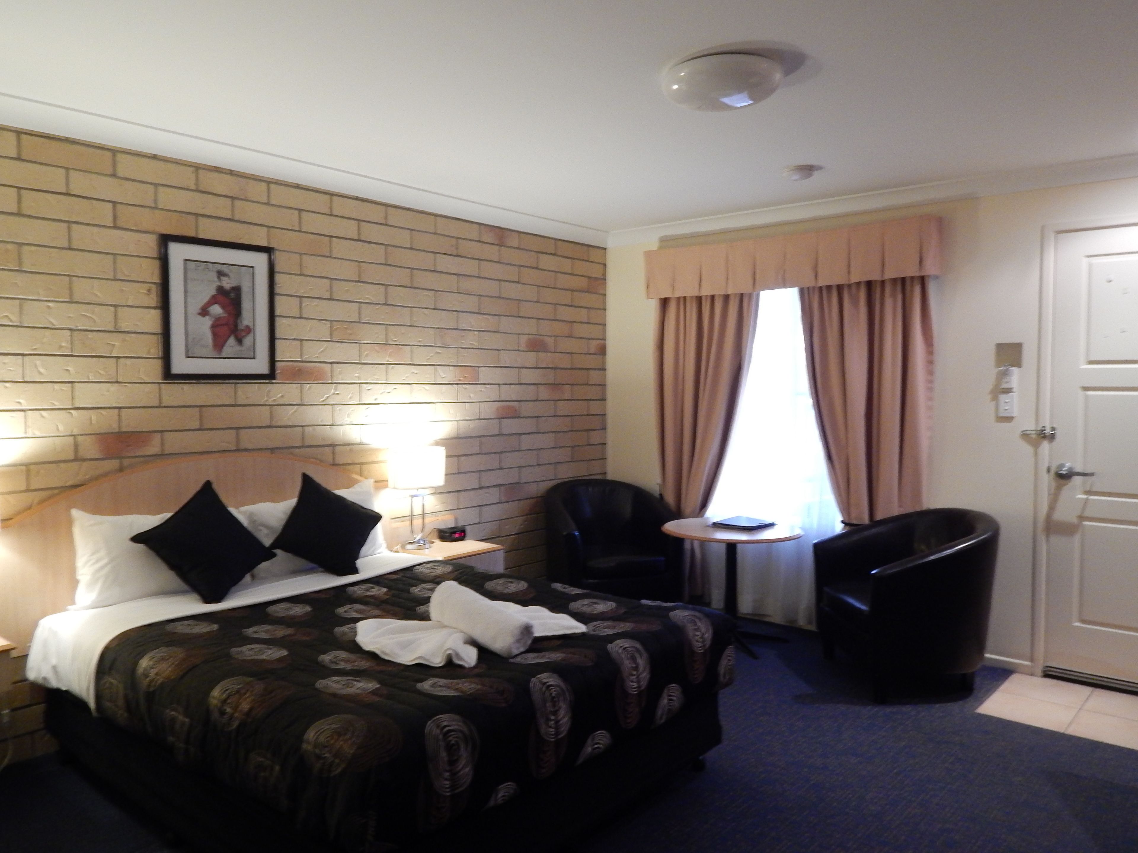 Quarto de hotel com parede em padrão de tijolos, uma cama com colcha estampada, uma mesa redonda pequena com uma cadeira, um cesto de lixo preto e uma porta.