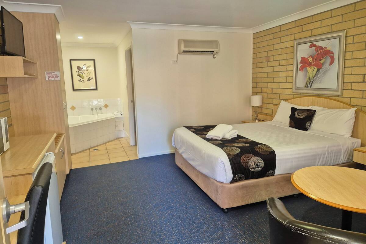 A hotel room with a bed, a desk, a chair, a round table, a bathroom with a bathtub, and a wall-mounted air conditioner.