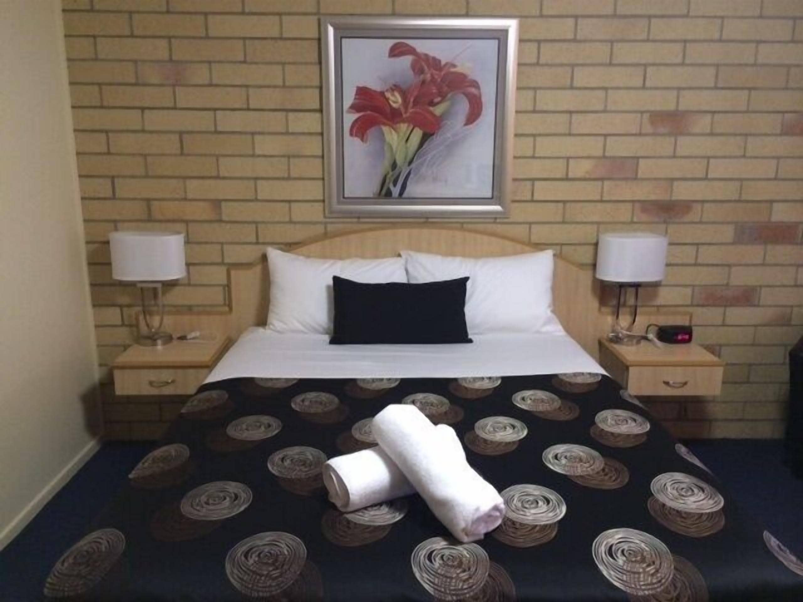 Een hotelkamer met een bakstenen muur, een bed met wit beddengoed, twee nachtkastjes met lampen en een lijst met een bloemenafbeelding aan de muur.