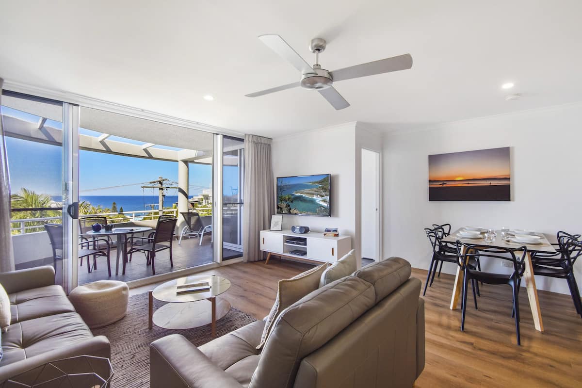 A modern living room with a large sliding glass door leading to a balcony with a view of the ocean. There is a ceiling fan, a flat-screen TV mounted on the wall, and a dining area with a table and chairs.