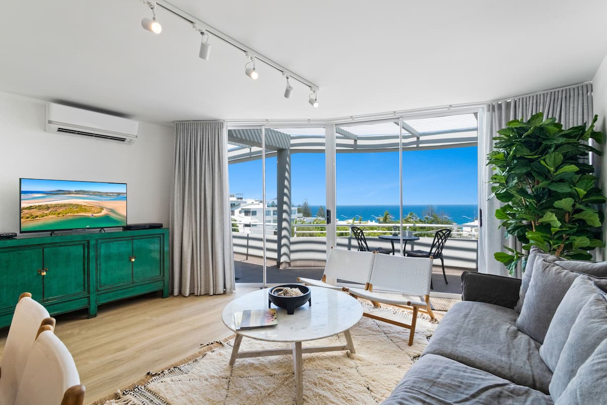 A modern living room with a large TV, a green cabinet, a round coffee table, and a view of the ocean through large windows.