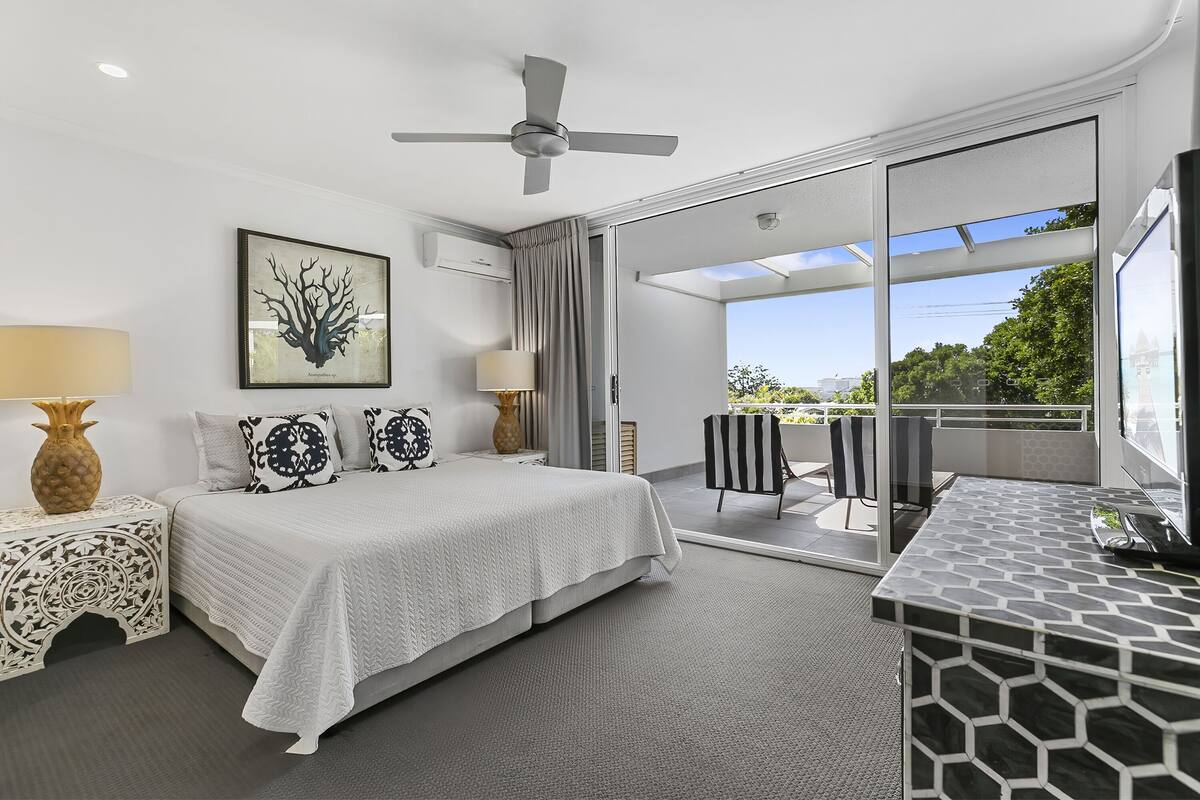 A modern hotel room with a bed, a desk, a chair, a ceiling fan, and a large window offering a view of the outdoors.