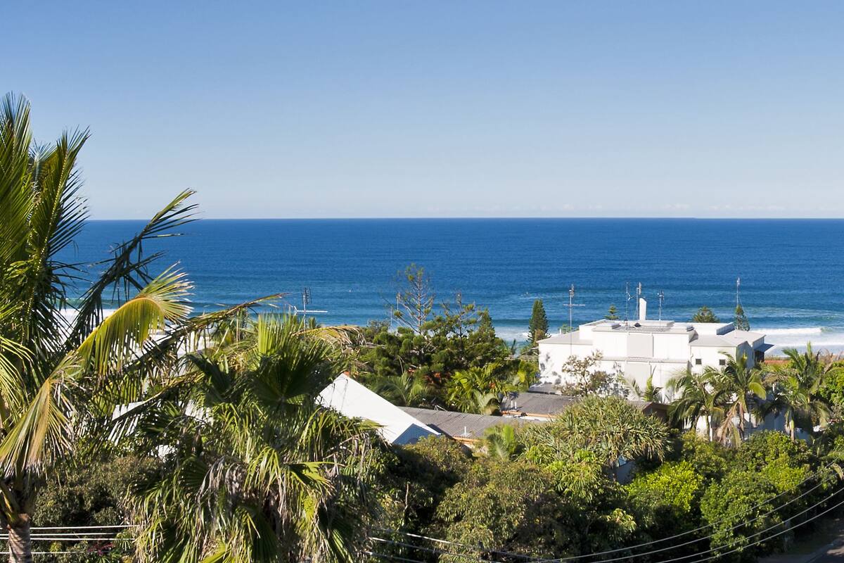 A coastal residential area with white buildings, a clear blue sky, and the ocean in the background.