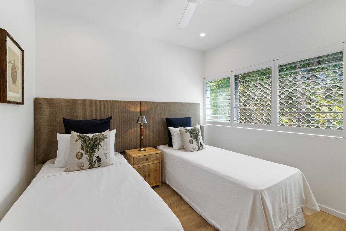 A bedroom with two beds, a wooden headboard, a nightstand with a lamp, and a window with a lattice screen.