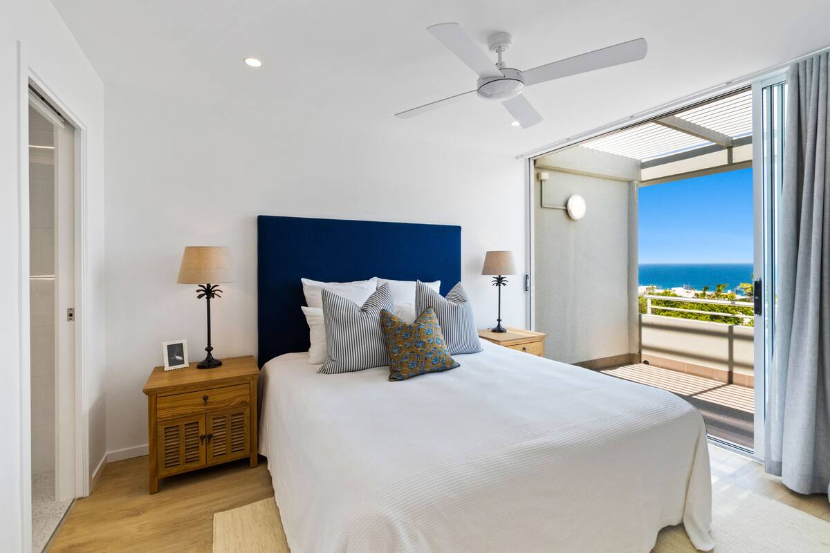 A bedroom with a large bed, a ceiling fan, and a view of the ocean through a balcony.