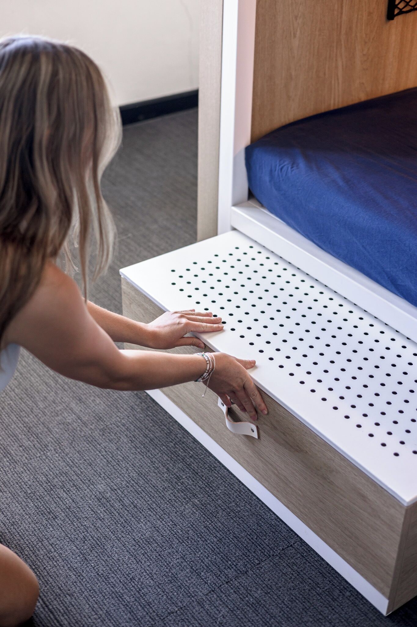 A person adjusting a white, perforated shelf in a room with a blue mattress and a wooden headboard.