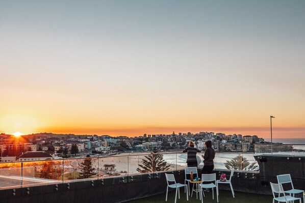 Une terrasse sur le toit offrant une vue sur le coucher de soleil au-dessus d’une ville, avec notamment une plage et des immeubles d’habitation.