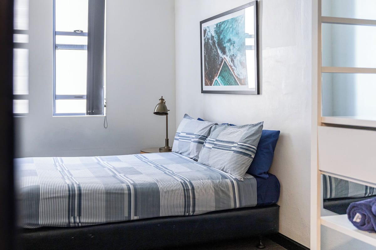 A bedroom with a bed, a nightstand, a lamp, a framed picture on the wall, and a window.