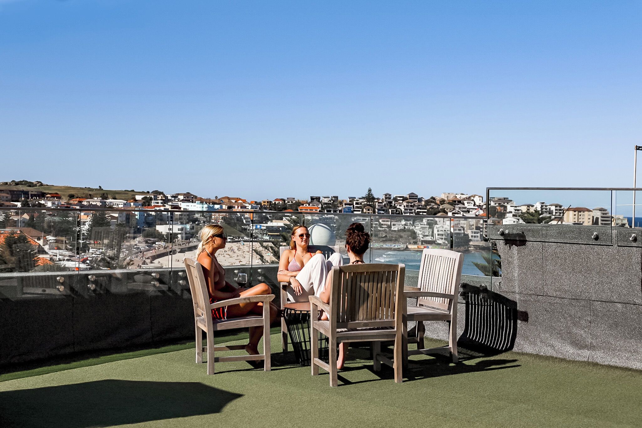 A rooftop terrace with three wooden chairs and a table, overlooking a coastal town.