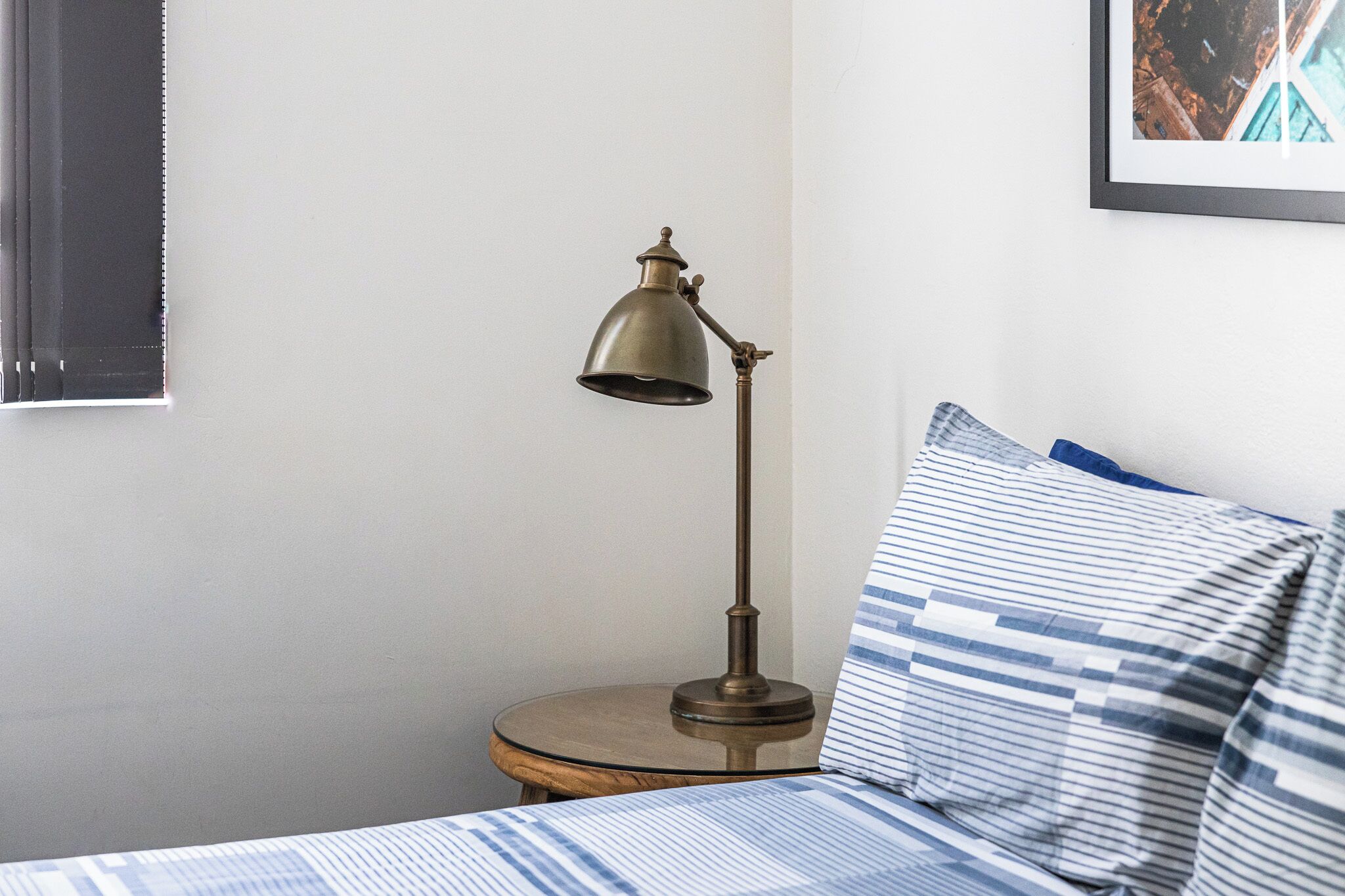 A bed with striped bedding, a round wooden nightstand, and a vintage-style desk lamp.