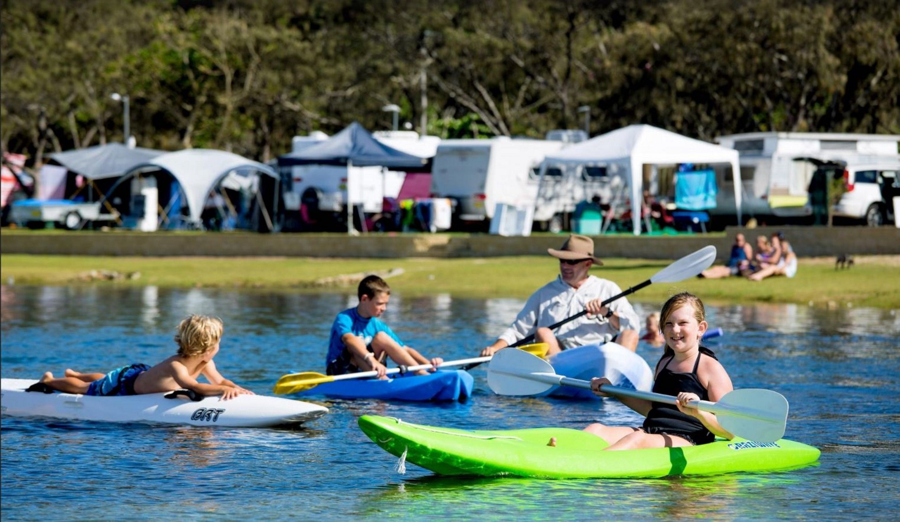Personas haciendo kayak en aguas tranquilas, con tiendas de campaña y vehículos al fondo.