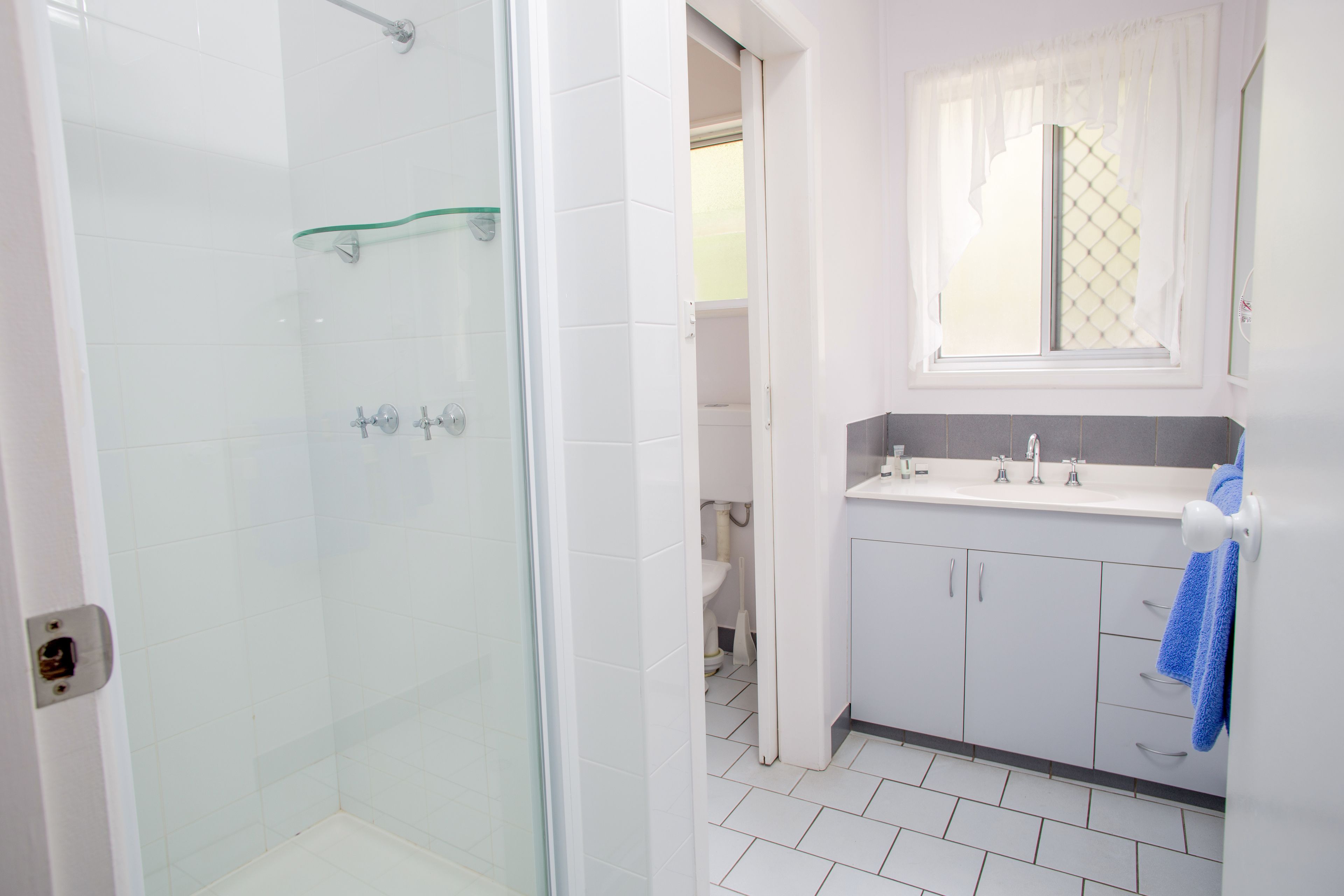 A bathroom with a glass shower enclosure, white tiled walls, a sink with a mirror, and a blue towel hanging on the wall.
