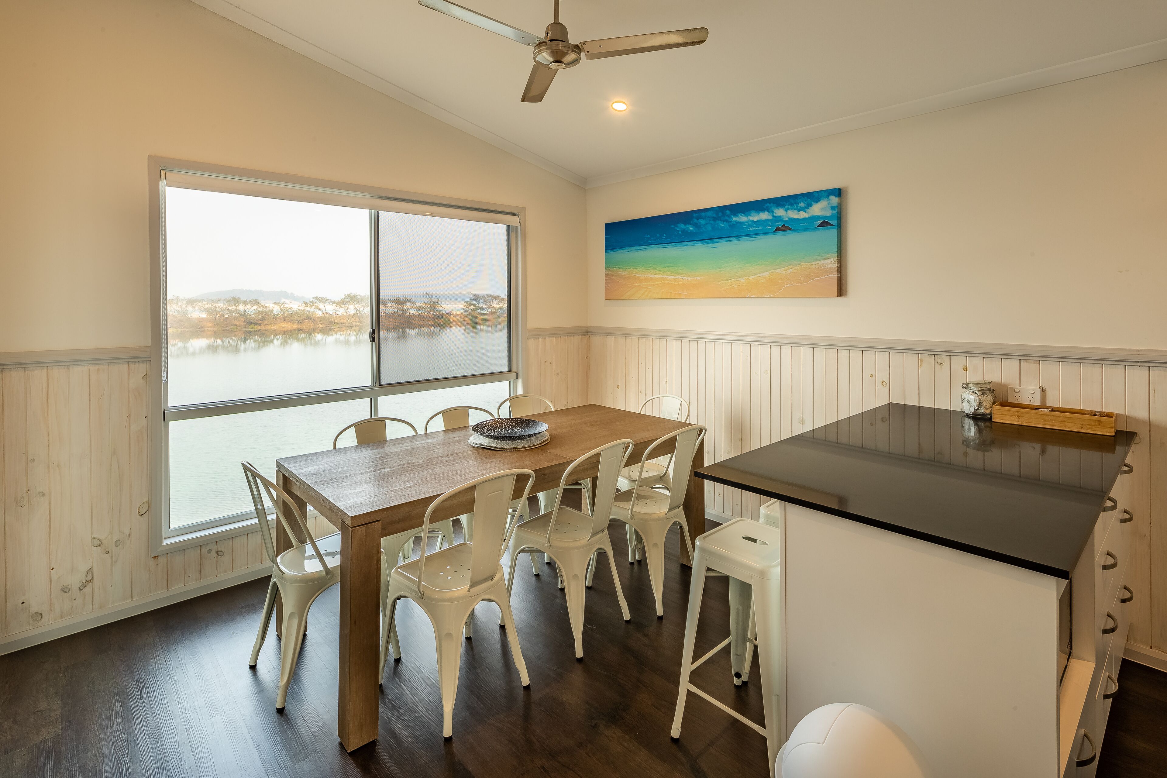A dining area with a wooden table and white chairs, a black countertop, and a large window overlooking a body of water.