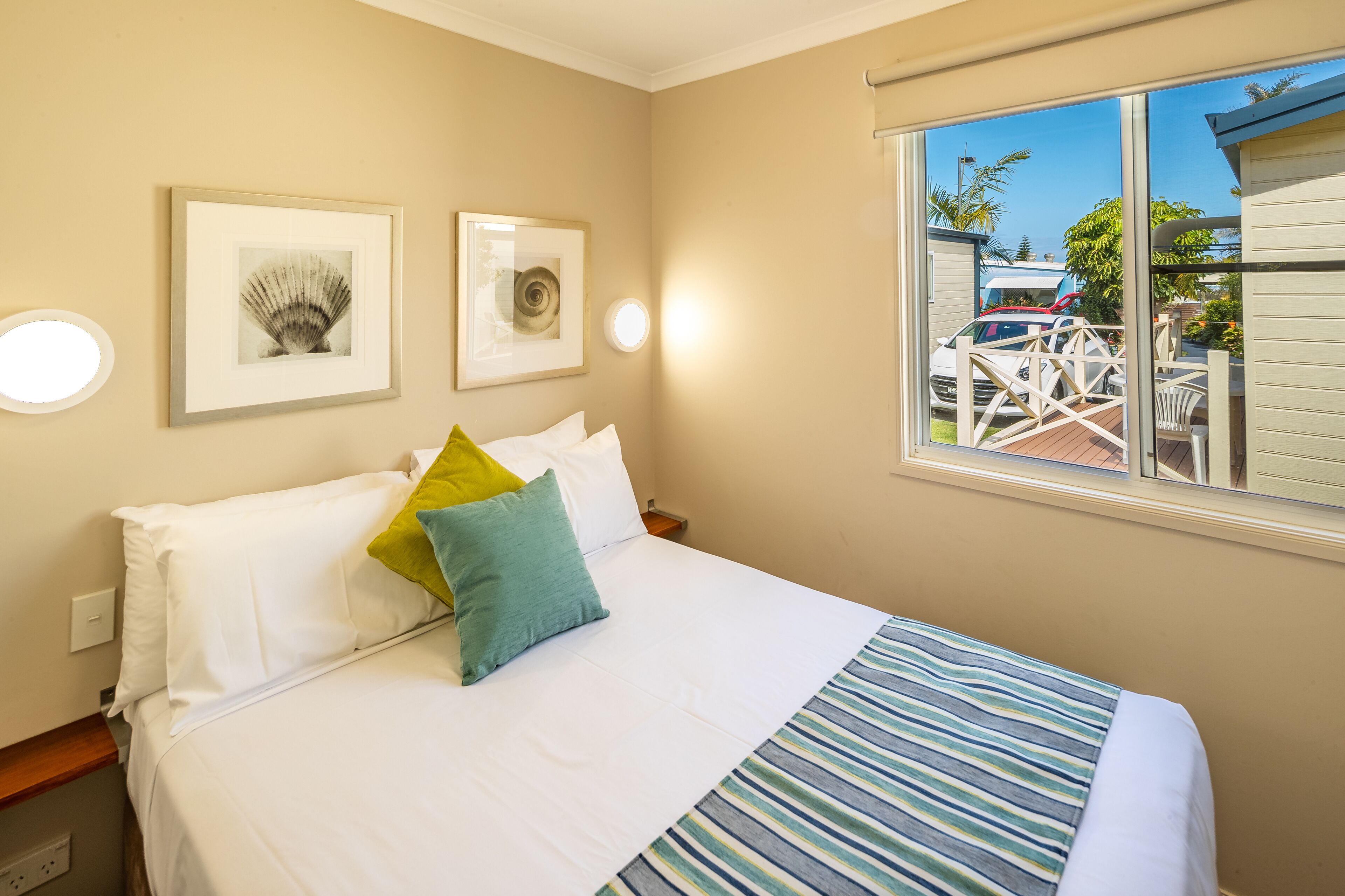 A neatly made bed with white and blue striped bedding, two framed pictures on the wall, and a window with a view of parked cars and greenery outside.