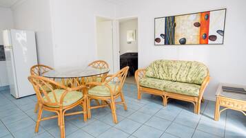 A room with a dining table surrounded by wicker chairs, a sofa, and a refrigerator.