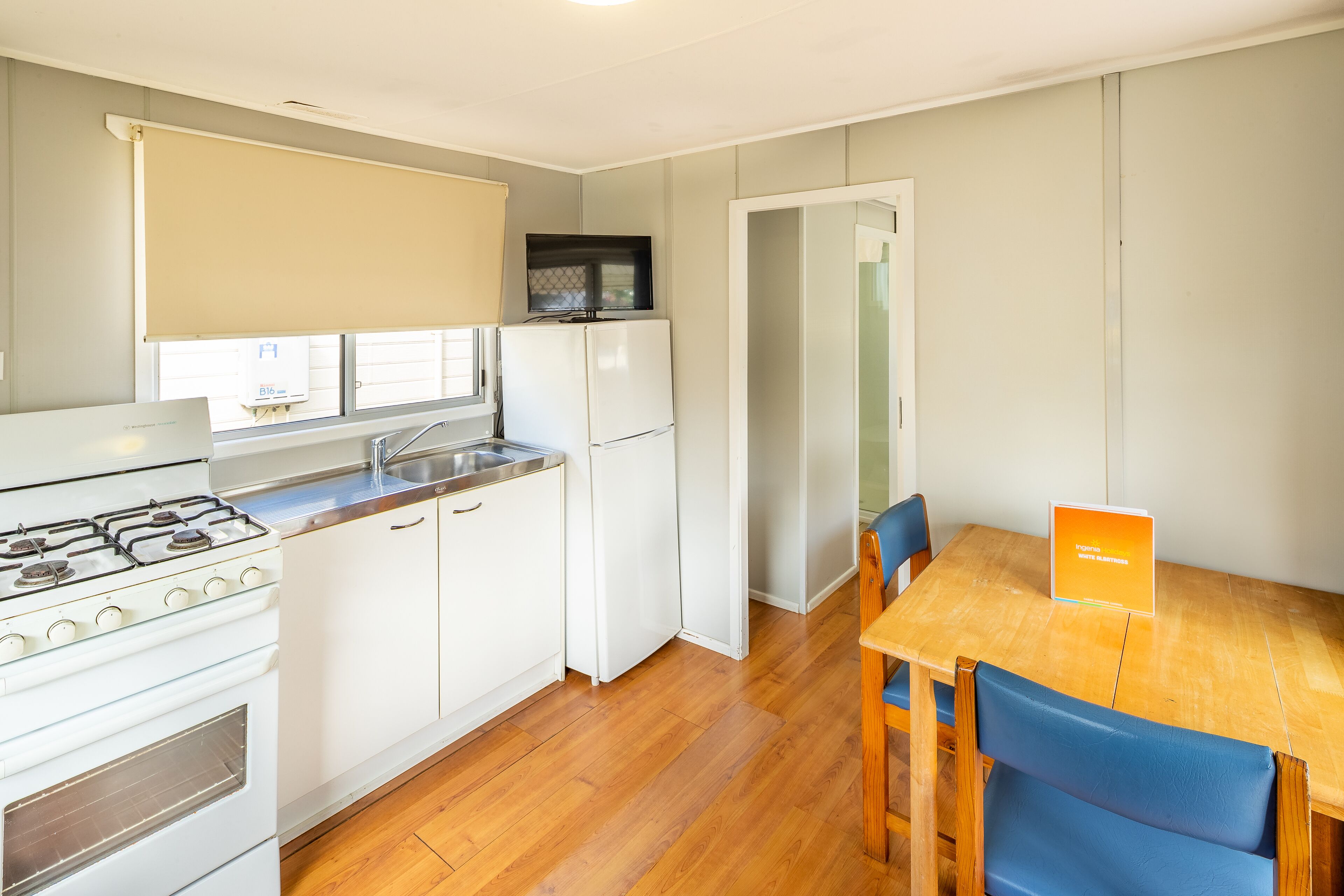 Cocina compacta con estufa, lavamanos y refrigerador, una mesa de comedor con sillas y vista a la calle a través de una ventana.