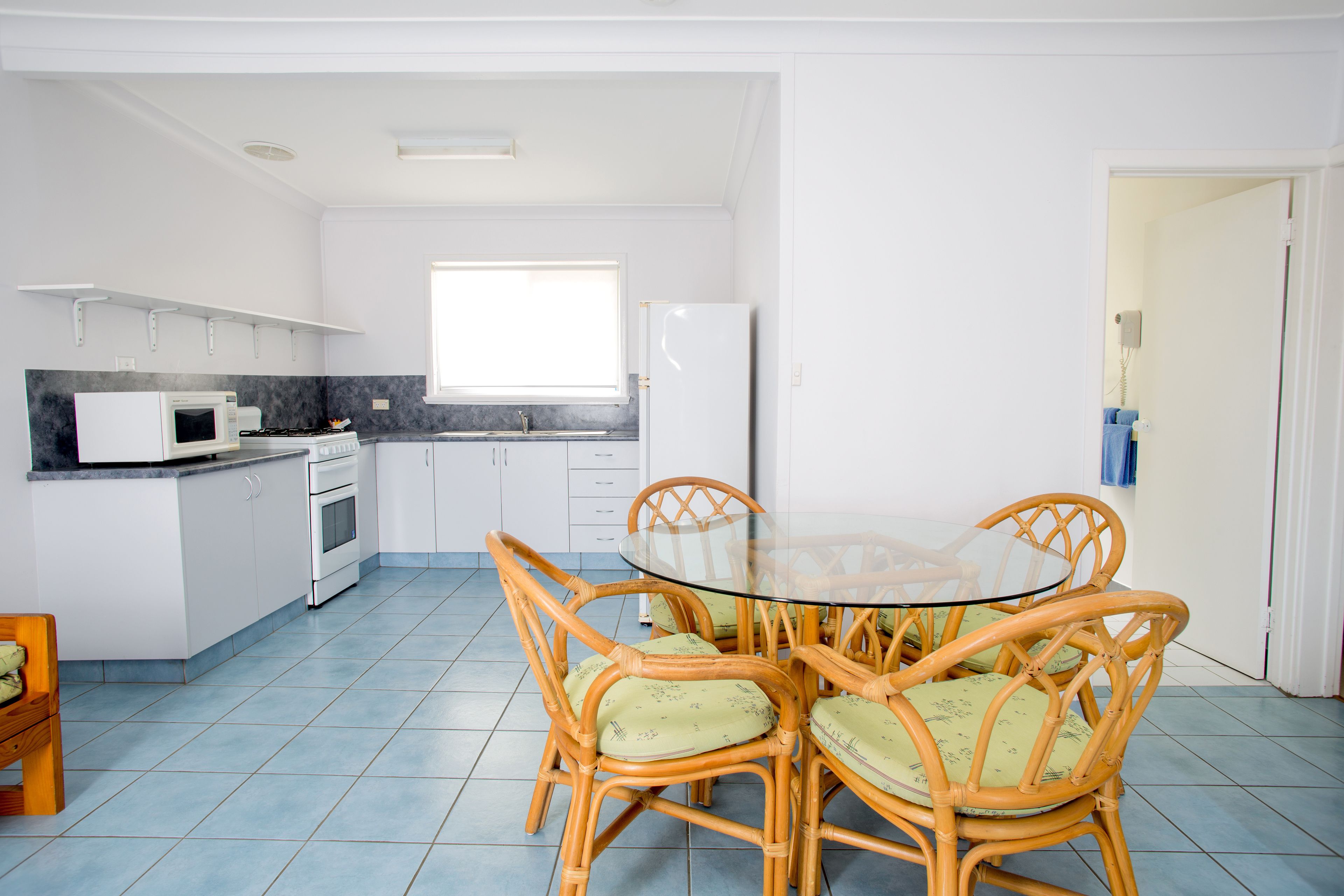 Cocina pequeña con gabinetes blancos, microondas, estufa y refrigerador. Hay un comedor con mesa de vidrio y sillas de mimbre.