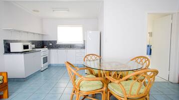 Cocina pequeña con gabinetes blancos, microondas, estufa y refrigerador. Hay un comedor con mesa de vidrio y sillas de mimbre.