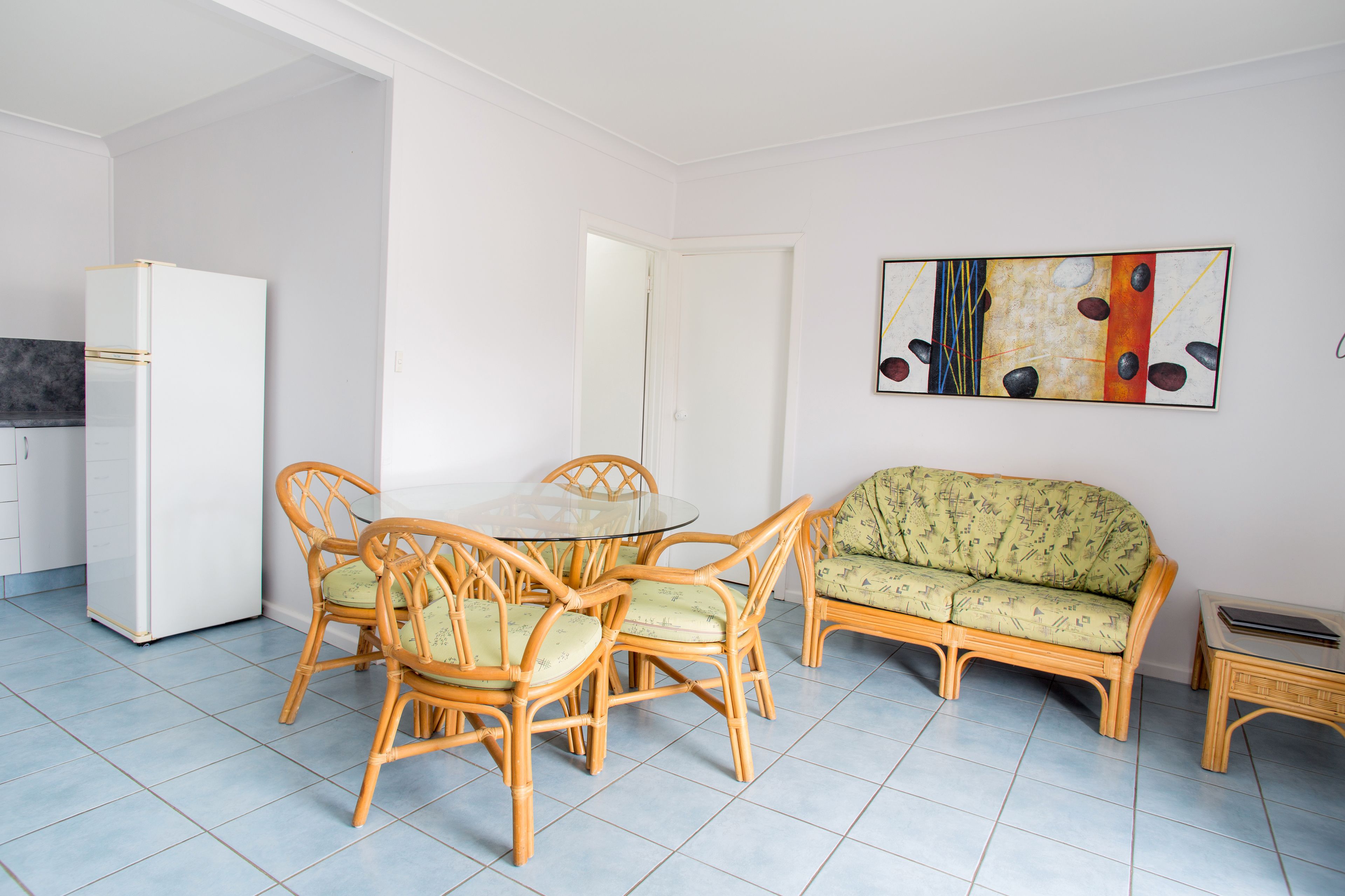 Un comedor con una mesa redonda de vidrio y cuatro sillas, un sofá verde y un refrigerador blanco.