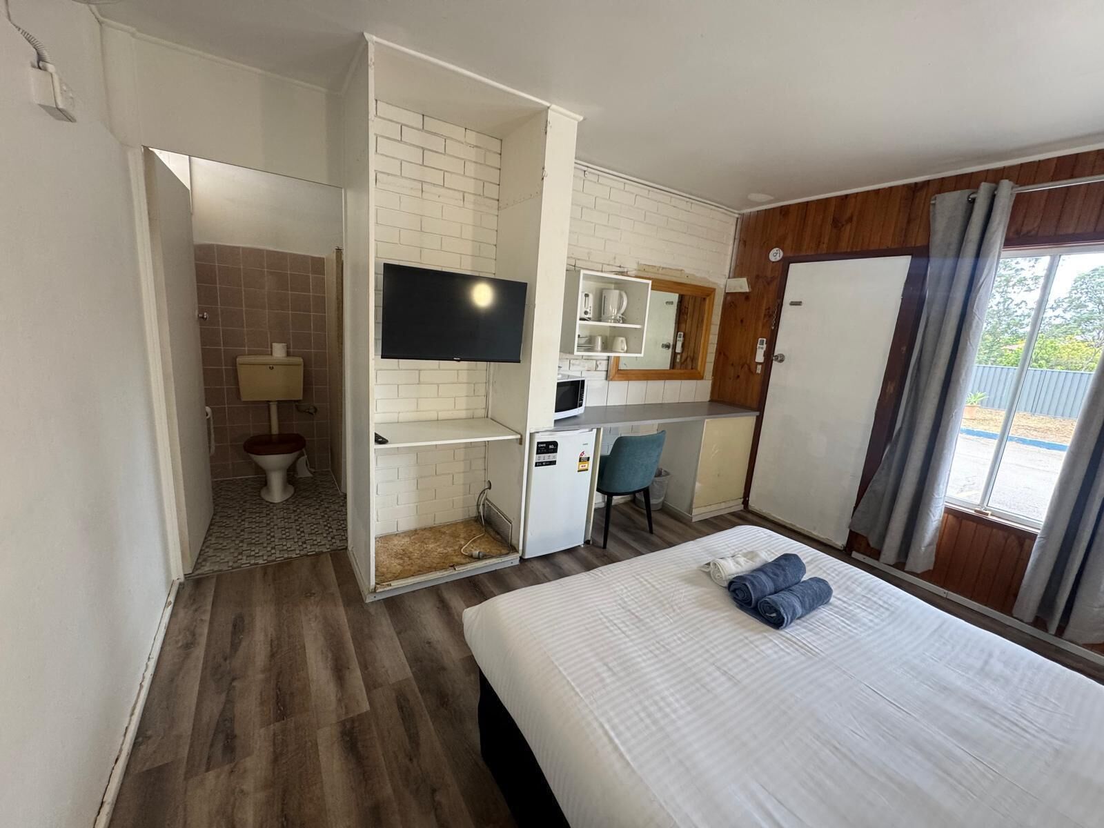A compact hotel room with a bed, a TV, a small kitchenette, a bathroom, and a view through a window.