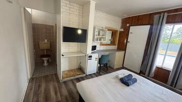 A compact hotel room with a bed, a TV, a small kitchenette, a bathroom, and a view through a window.