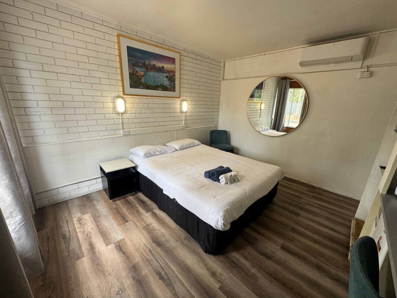 A hotel room with a bed, a bedside table, a chair, a round mirror, and a wall-mounted picture.