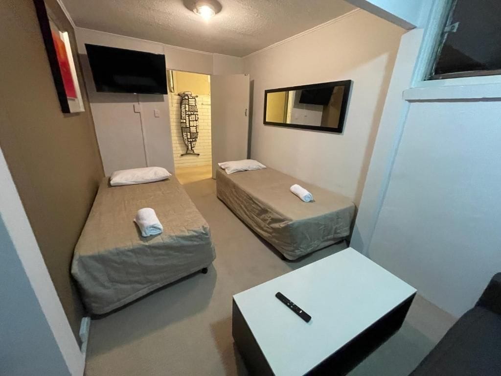 A small, compact room with two single beds, a flat-screen TV mounted on the wall, a small table, and a window.