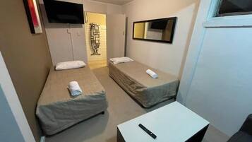 A small, compact room with two single beds, a flat-screen TV mounted on the wall, a small table, and a window.