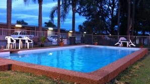 Una piscina rectangular con borde de azulejos, rodeada por un bajo muro de ladrillo. Hay dos sillas de plástico blancas y una mesita cerca de la piscina, y una valla con un coche blanco estacionado detrás.