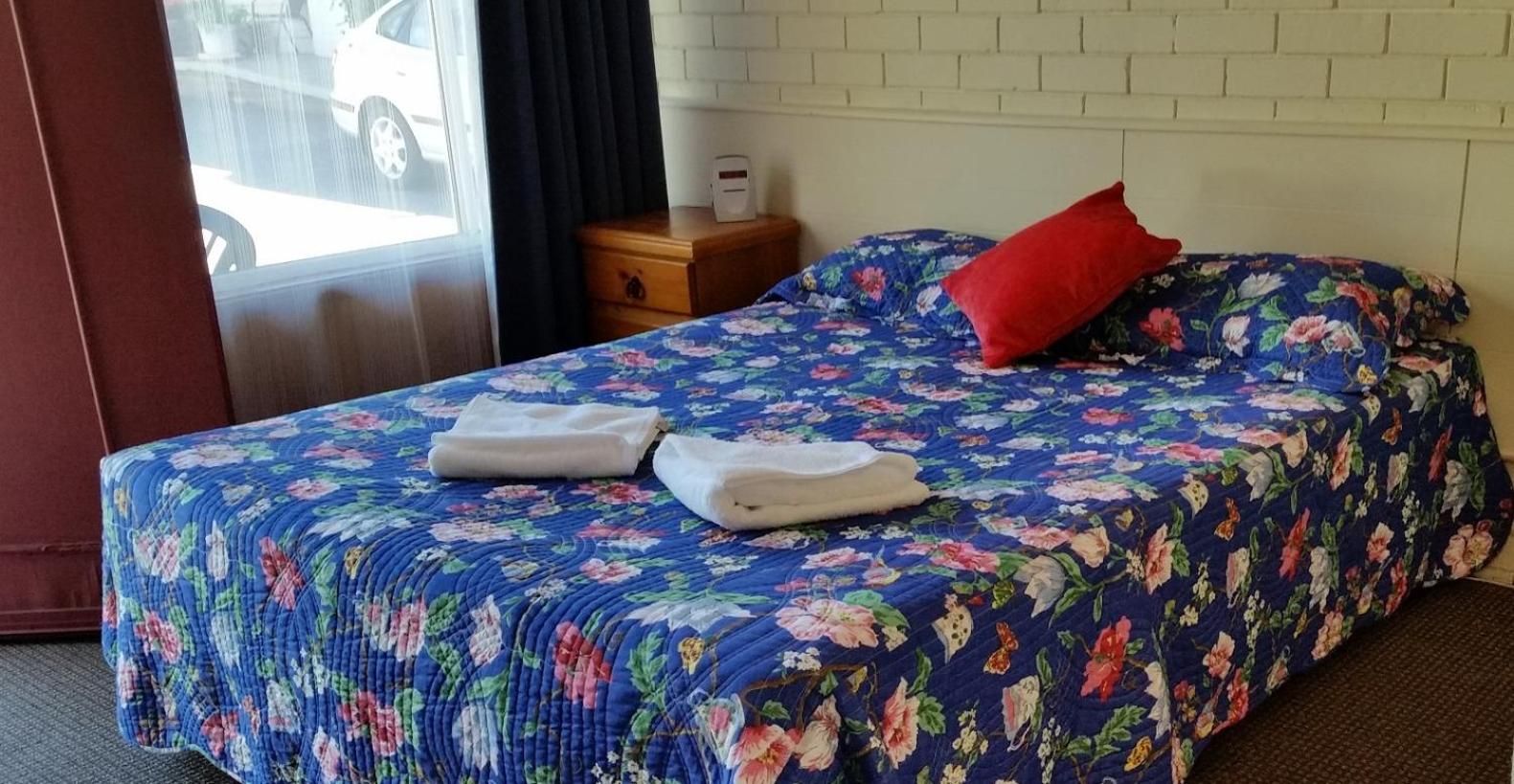 A hotel room with a floral bedspread, a red pillow, and two folded towels on the bed.