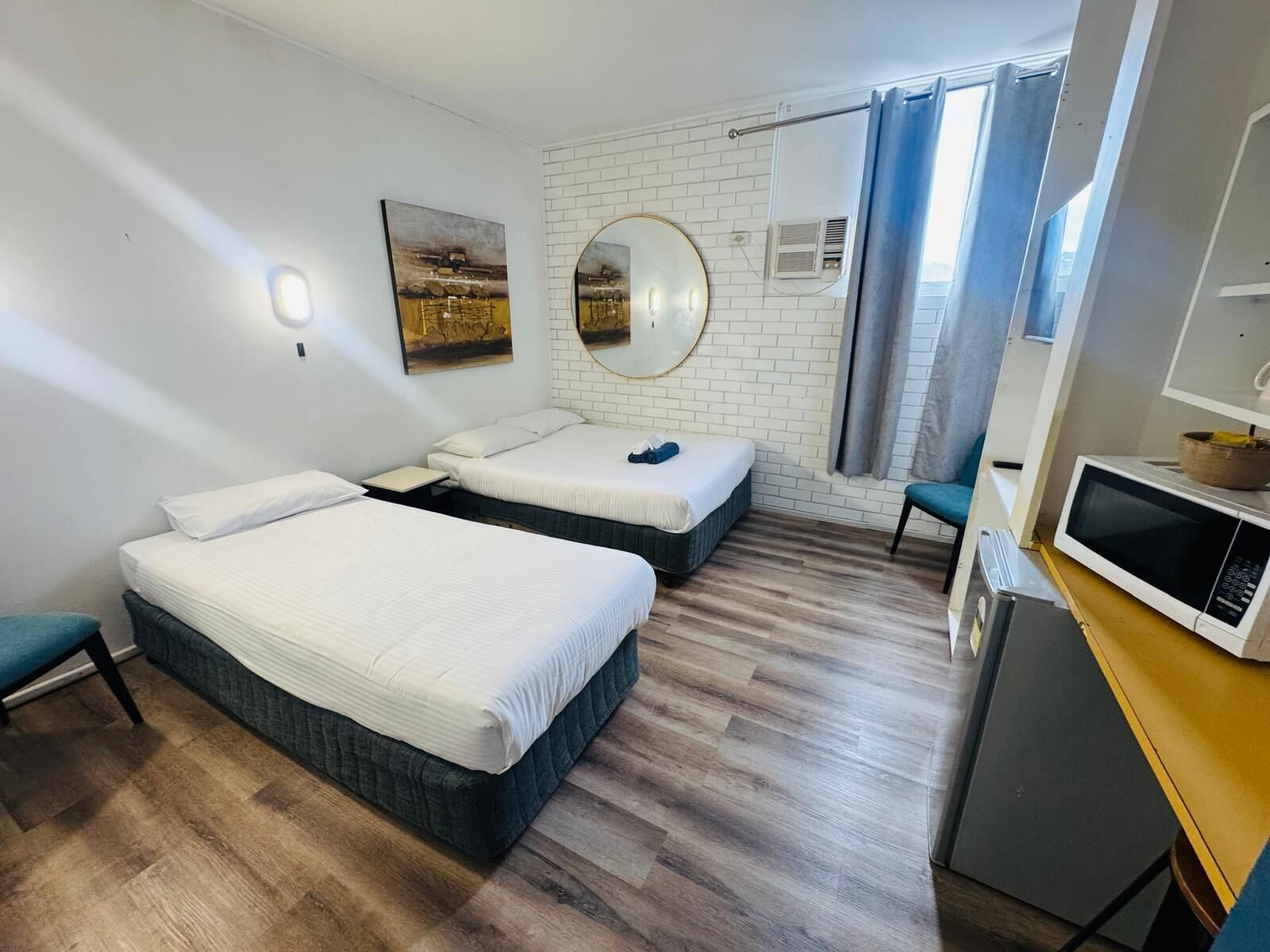 Ein Hotelzimmer mit zwei Betten, einem Spiegel, einem kleinen Schreibtisch mit einer Mikrowelle und einem Fenster mit Vorhängen.
