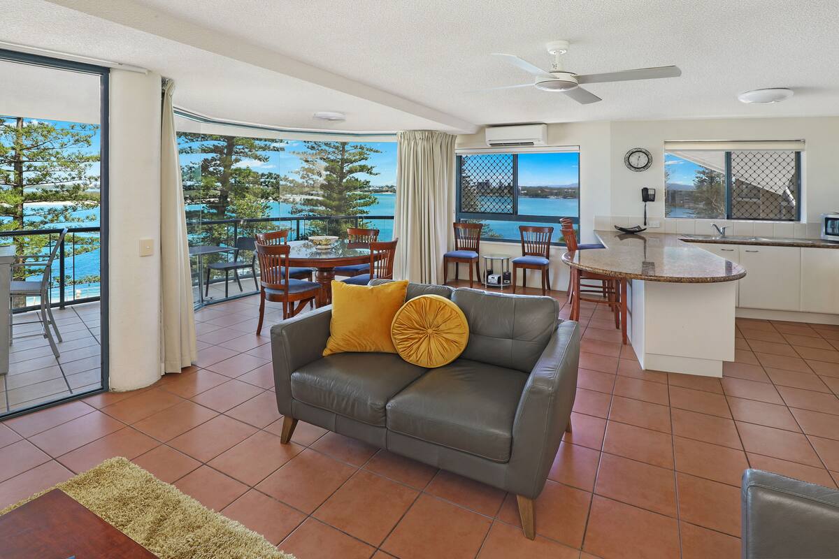 Amplia sala de estar con un sofá de cuero gris, sillas de madera, una mesa de comedor y una zona de cocina. Un ventanal ofrece vistas al mar y a los árboles.