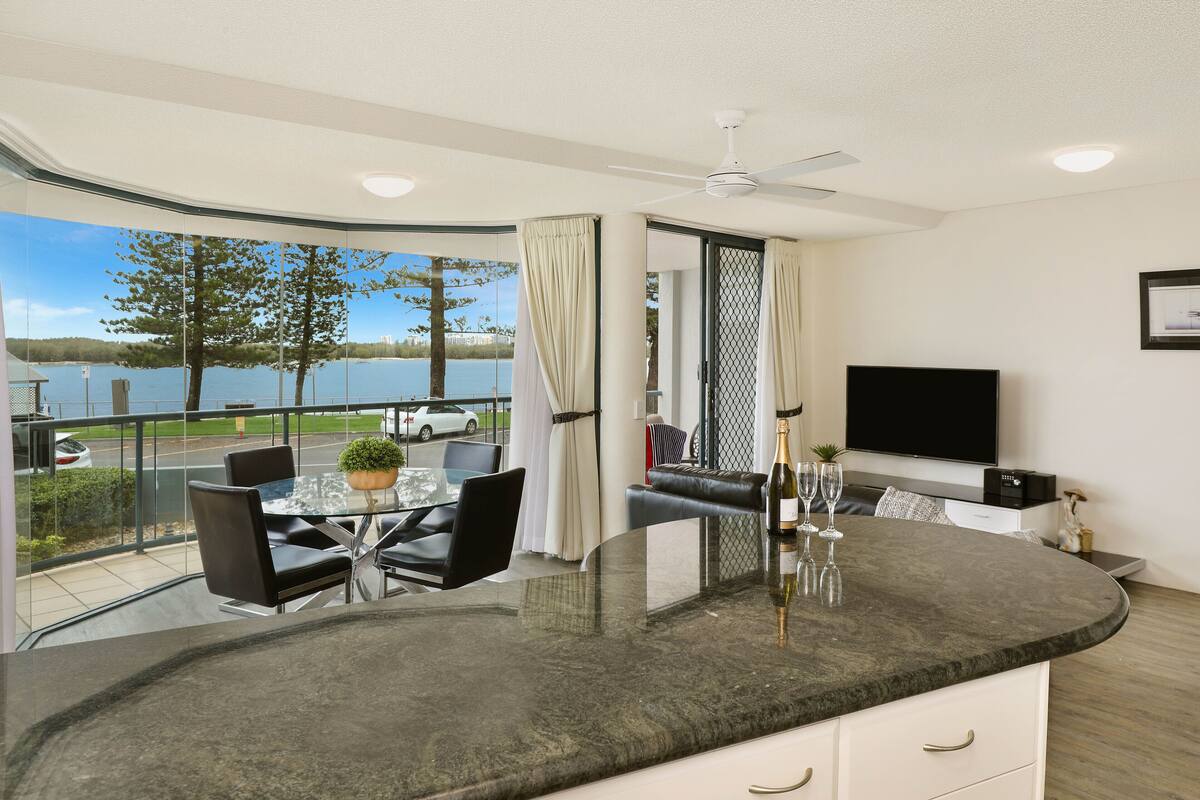 Una cocina moderna con una isla curva, una zona de comedor con mesa y sillas de vidrio, un televisor y vistas a un lago.