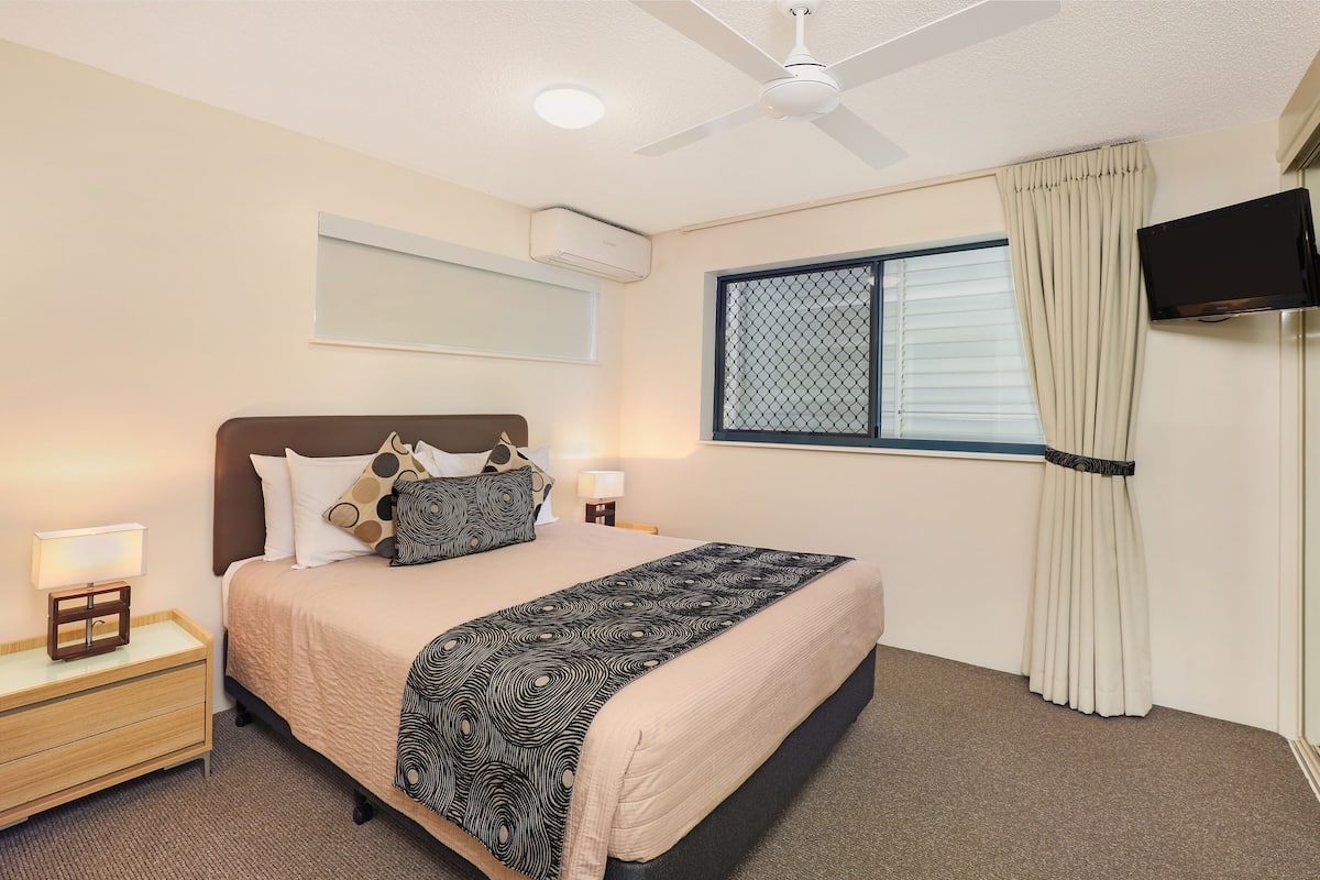 Habitación de hotel con cama, mesitas de noche, ventilador de techo, unidad de aire acondicionado, ventana con cortinas y televisor montado en la pared.