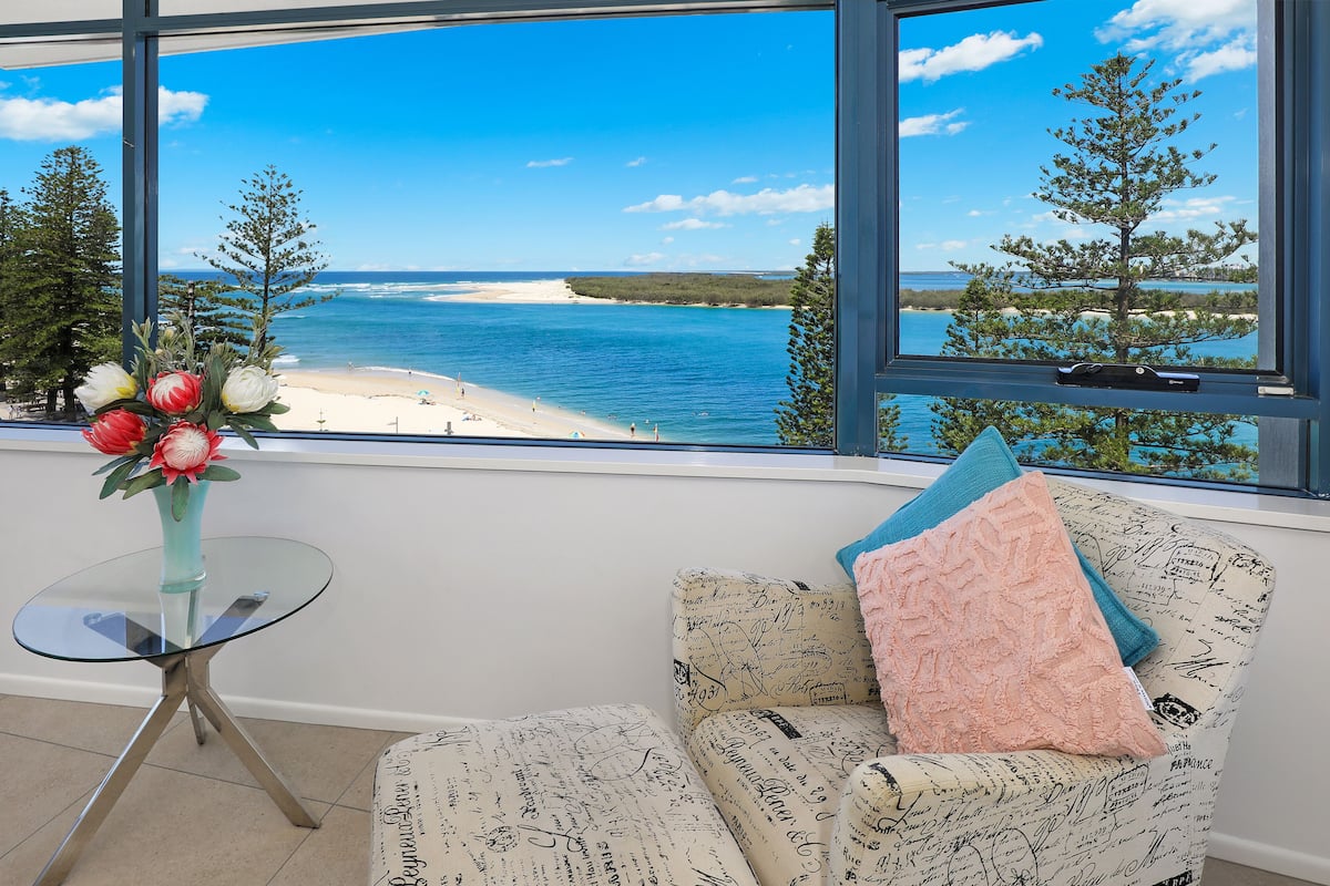 Una habitación con un ventanal grande que ofrece vista al mar, una mesa de vidrio con un jarrón de flores y un sillón estampado.