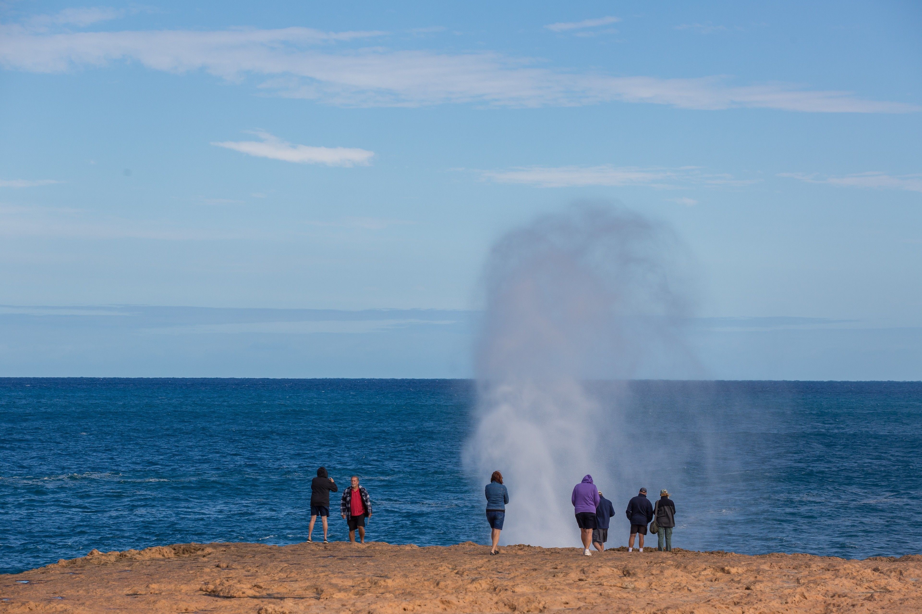 Un grupo de personas de pie en una orilla rocosa junto al mar, observando un géiser.