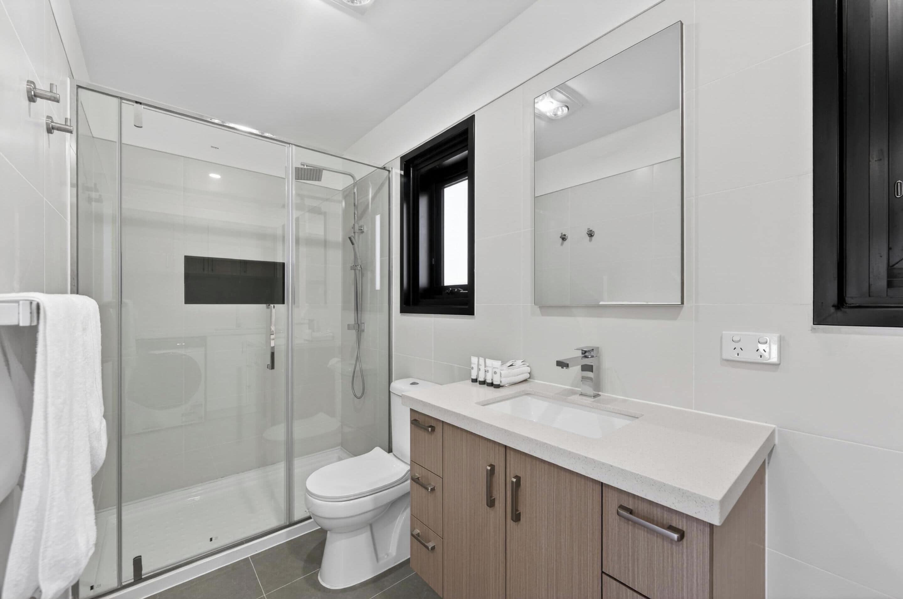 A modern bathroom with a glass shower enclosure, a white toilet, a sink with a mirror above, and wooden cabinets.