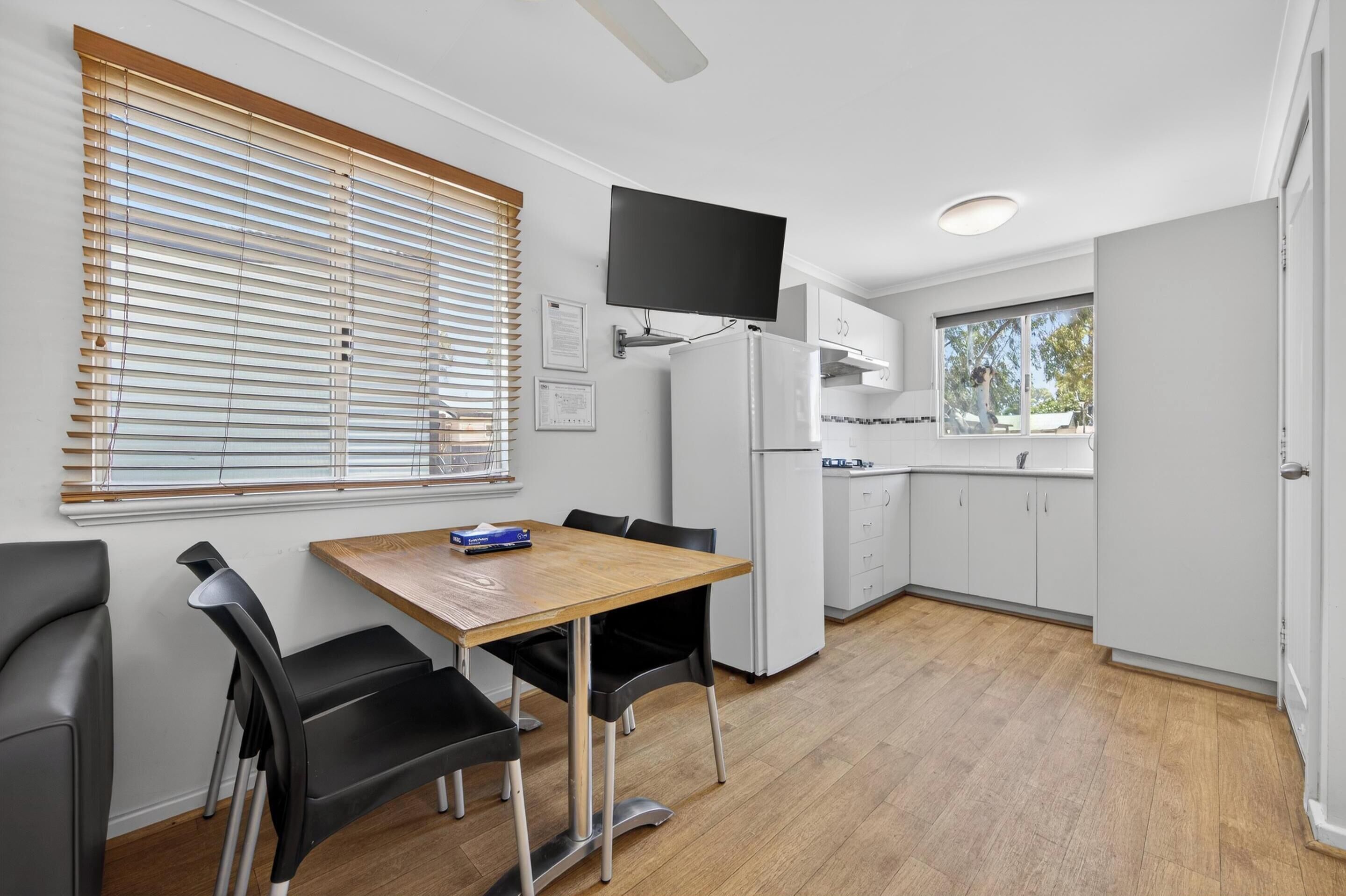 Una cocina moderna con zona de comedor, armarios blancos, televisor de pantalla plana y una ventana con persianas.