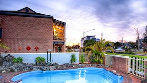A swimming pool with a brick border, a building with a balcony, and a street view in the background.