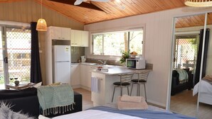 A compact hotel room with a kitchenette, a bed with a blue and white striped bedspread, a small dining area with a bar stool, and a view through a sliding door.
