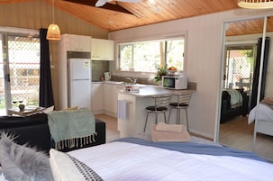 Quarto de hotel compacto com cozinha compacta, uma cama com colcha listrada em azul e branco, um pequeno espaço de refeições com um banqueta e vista através de uma porta de correr.