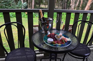 Uma mesa de vidro com uma garrafa de champanhe, duas taças de vinho e um prato com uvas, azeitonas e queijo sobre um deck de madeira com cadeiras.