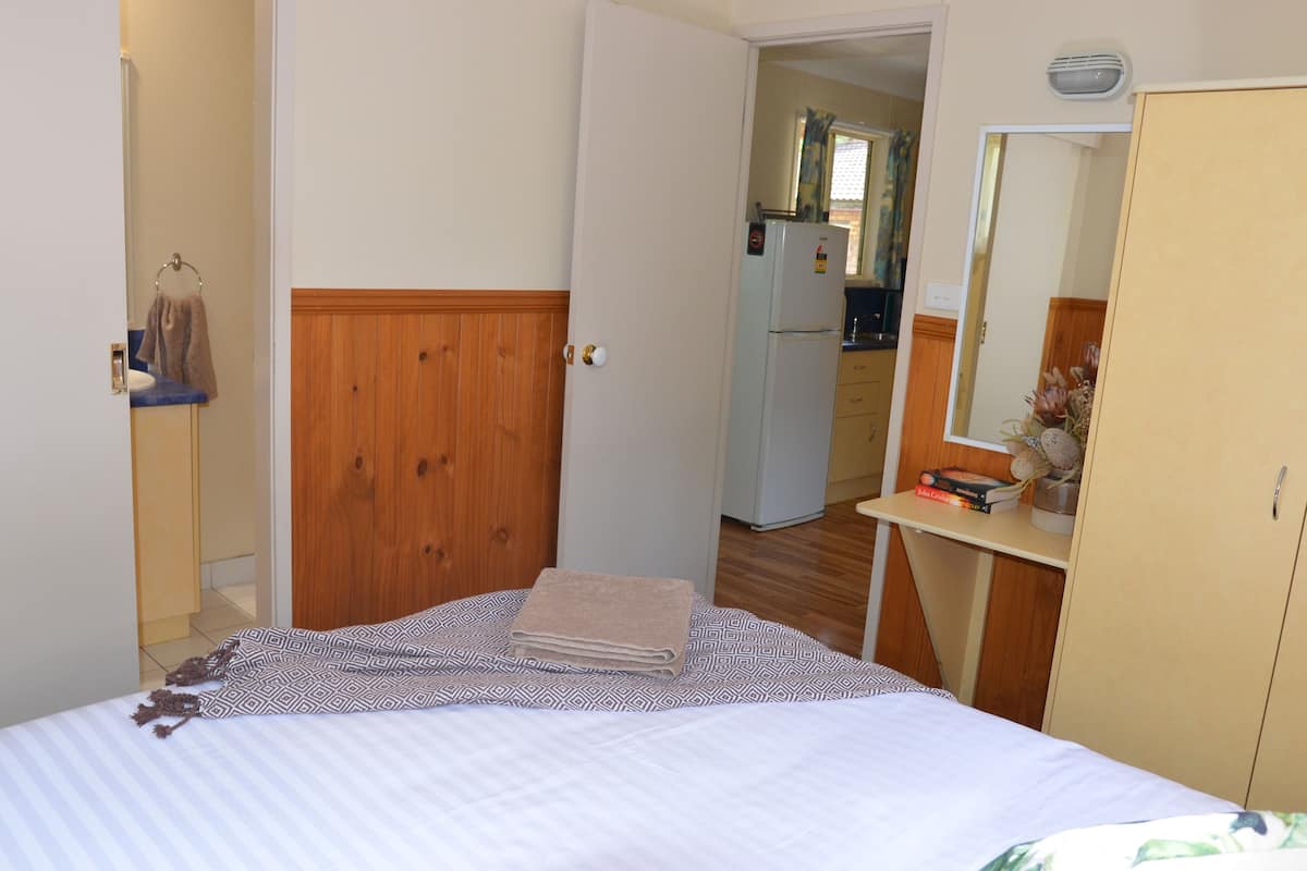 Un dormitorio con cama, un armario de madera, un pequeño escritorio con espejo y una kitchenette al fondo.