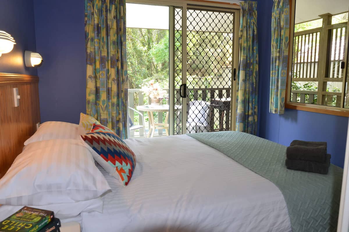 Un dormitorio con cama, almohadas, una ventana con cortinas, vistas al exterior y una pequeña mesa con sillas afuera.