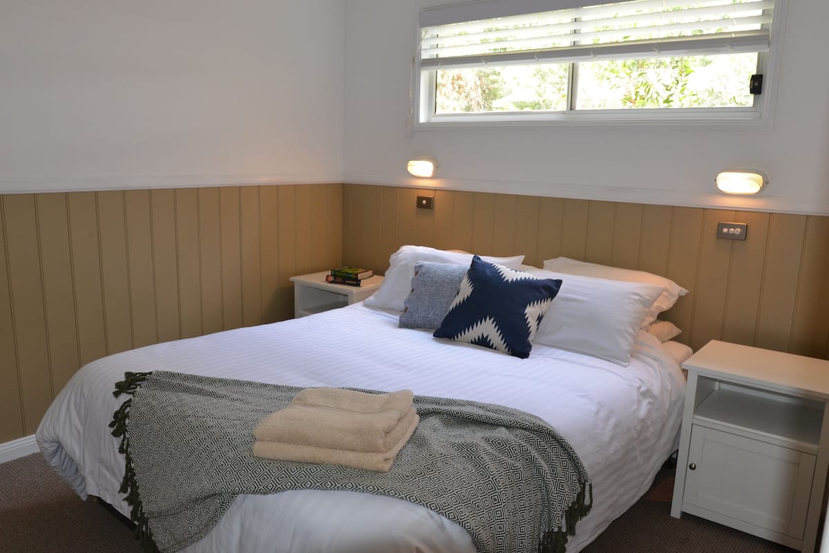 Una cama bien hecha con una manta y almohadas estampadas, una mesita de noche con una lámpara y una ventana con persianas.